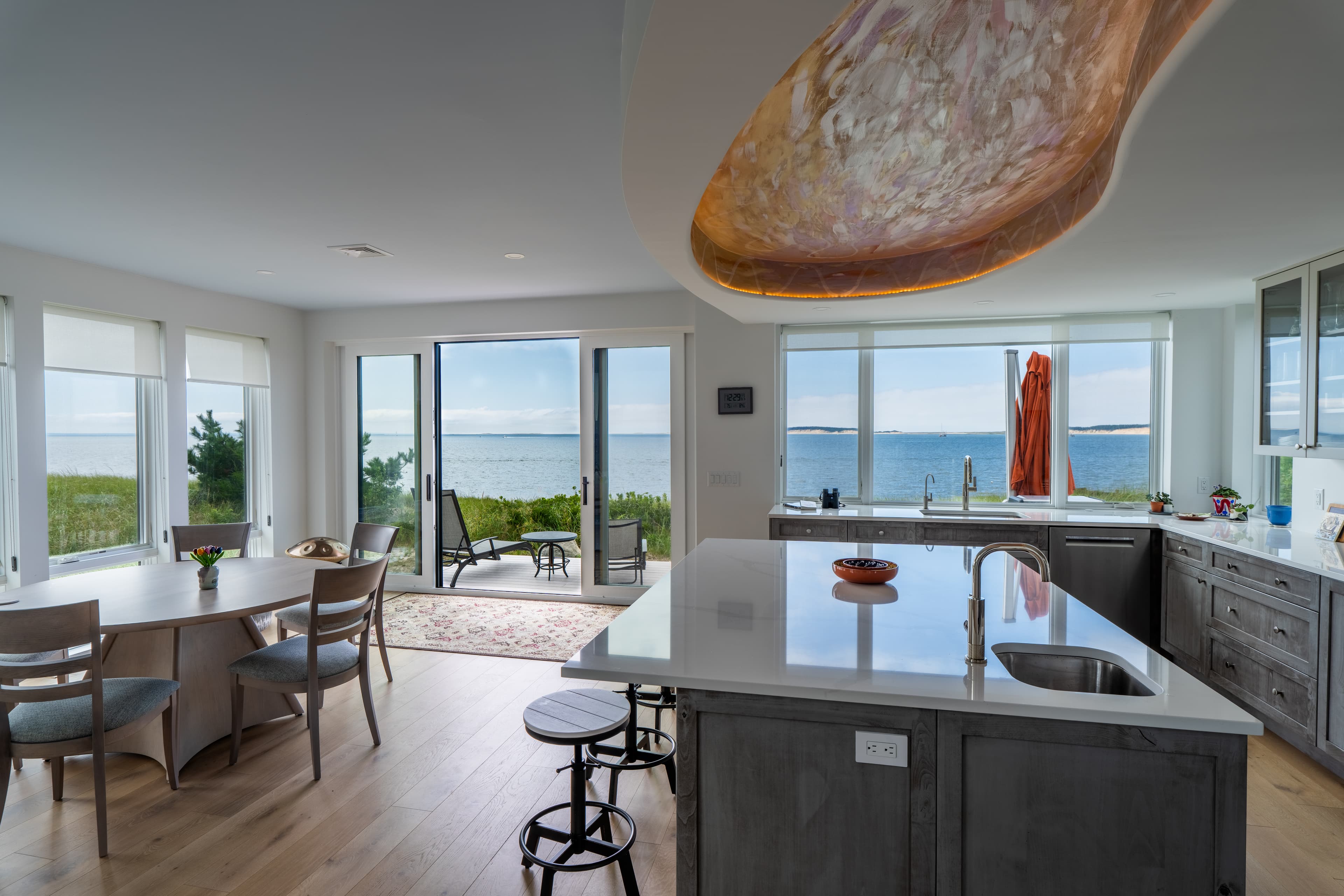 Gray-washed cabinetry, glass-front uppers and a white quartz island sit under the soffit, with the harbor filling the wall of glass beyond.